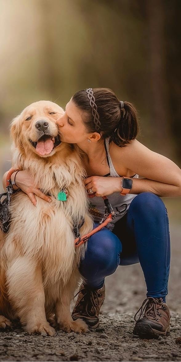 A woman kneels on a forest trail and kisses her smiling golden retriever on the cheek.